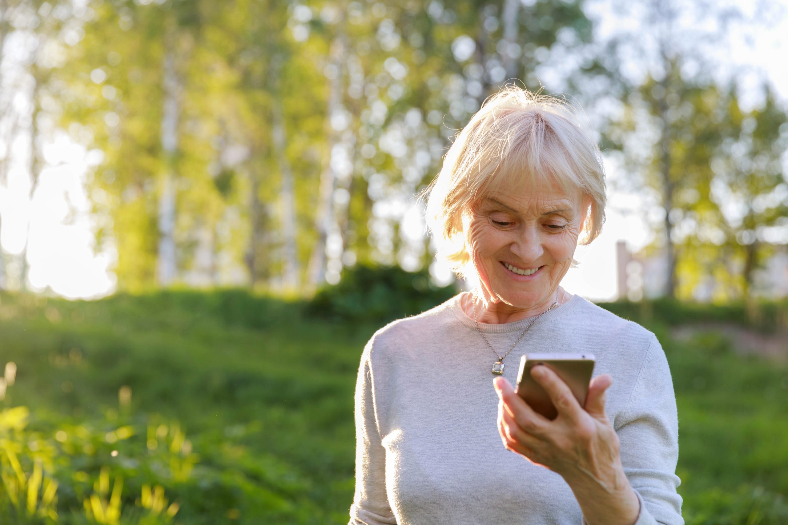 Elderly woman is holding a phone in a sunny park. Show care and