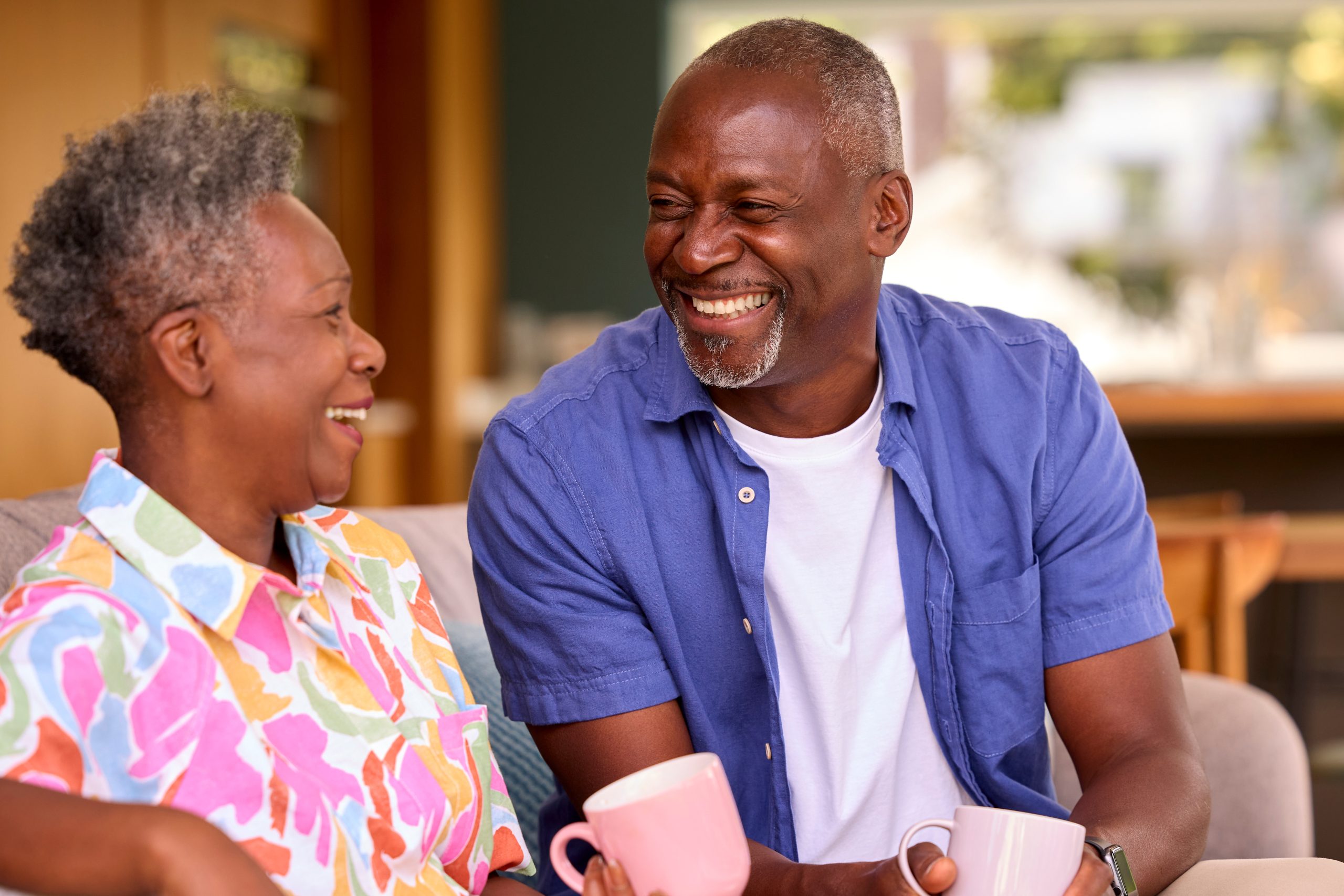 Smiling Senior Couple Sitting On Sofa At Home Drinking Coffee An